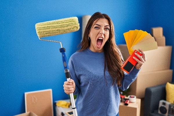 Young brunette woman holding roller painter painting new house angry and mad screaming frustrated and furious, shouting with anger. rage and aggressive concept.